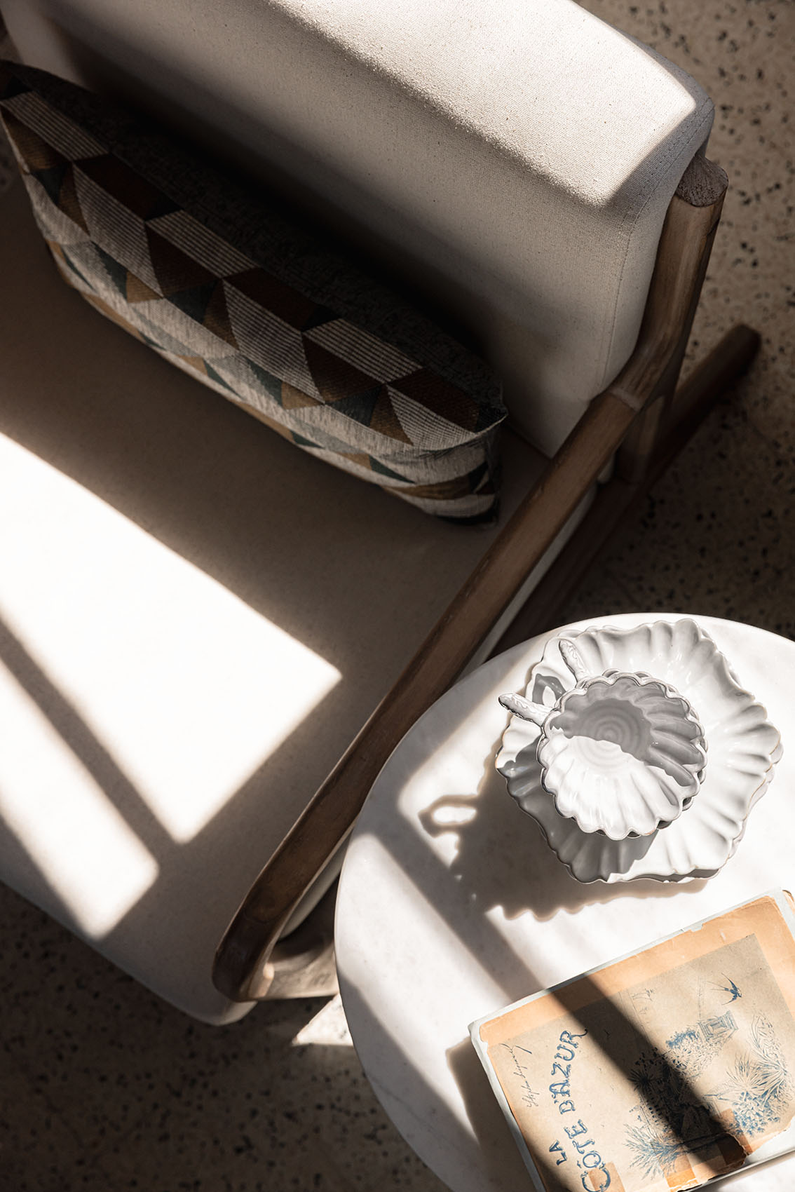 Armchair arm with geometric cushion, shell bowl, Côte d'Azur book and shadows on terrazzo