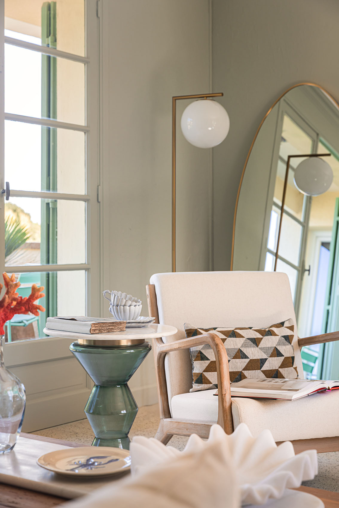 Armchair, green glass side table with books and shell cup, tall oval gold mirror and globe lamp