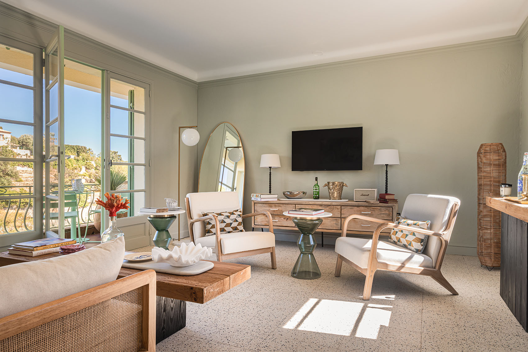 Sage-walled living room with wooden armchairs, chest of drawers, oval mirror and open French doors