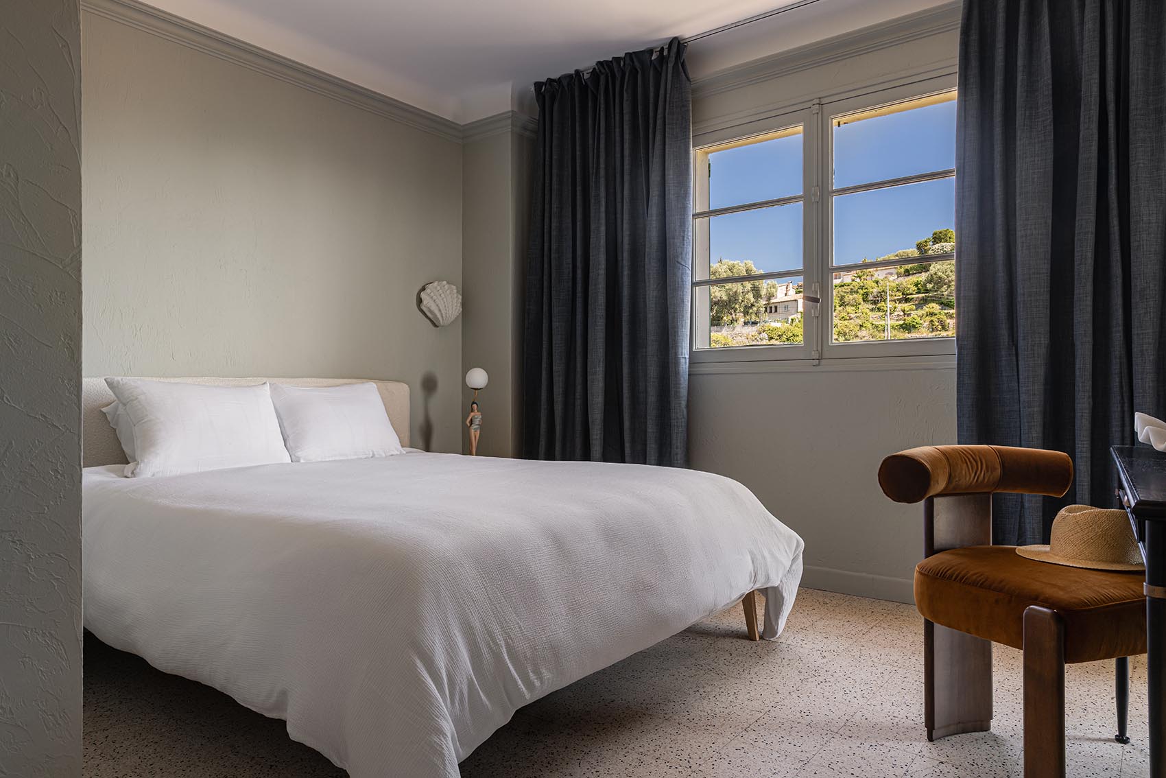 Bedroom with white bed, heavy grey drapes, hillside window and caramel armchair