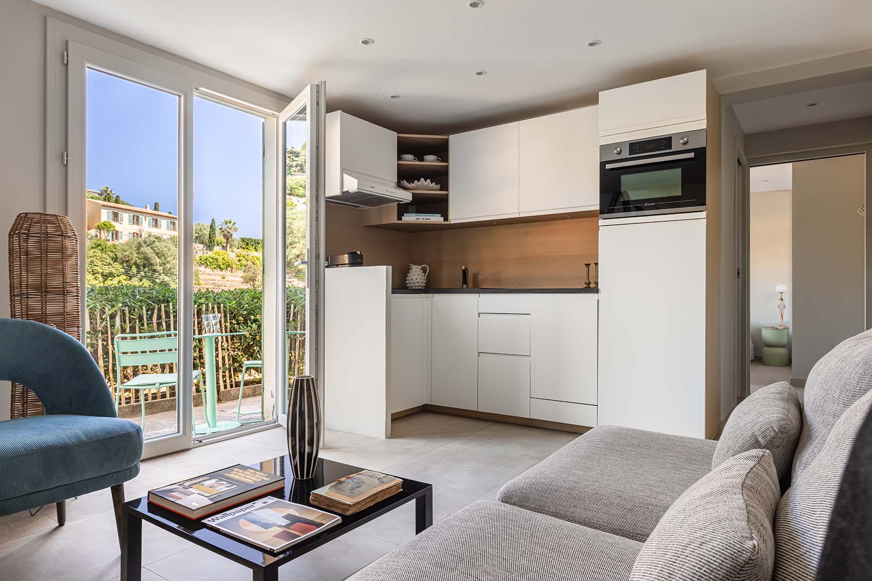Studio with white kitchen, grey sofa and blue velvet armchair facing French doors to the garden