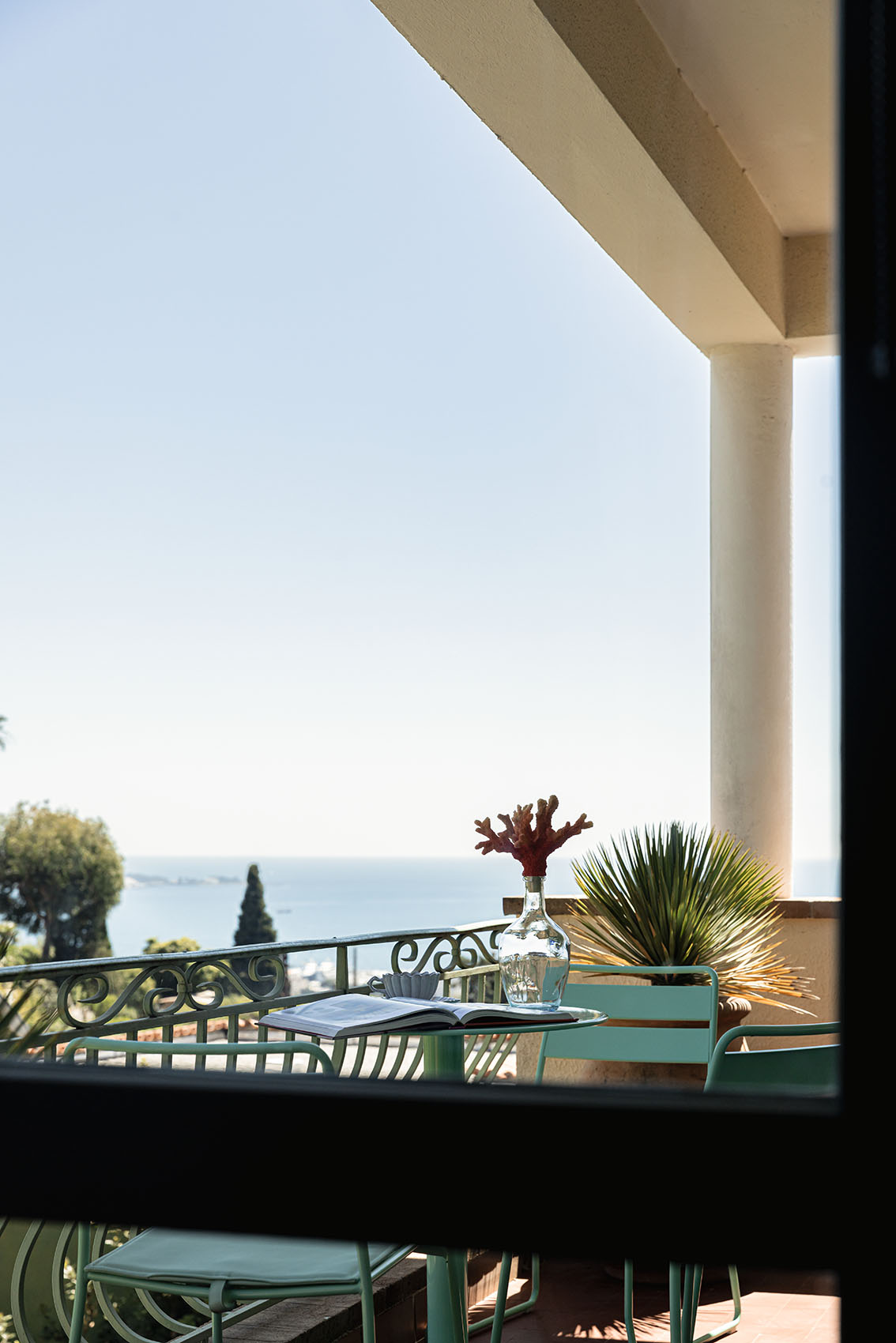 Balcony with green bistro table, coral in vase, open book and sea view between the pillars