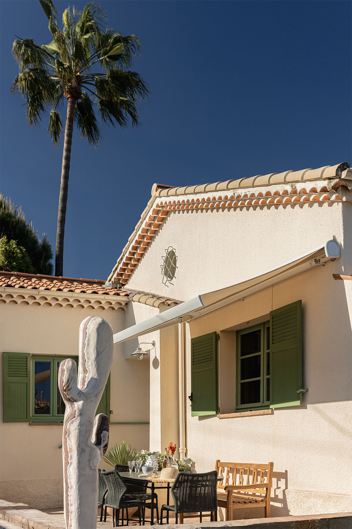 Provençal facade with round tiles, green shutters, cactus sculpture and palm tree under blue sky