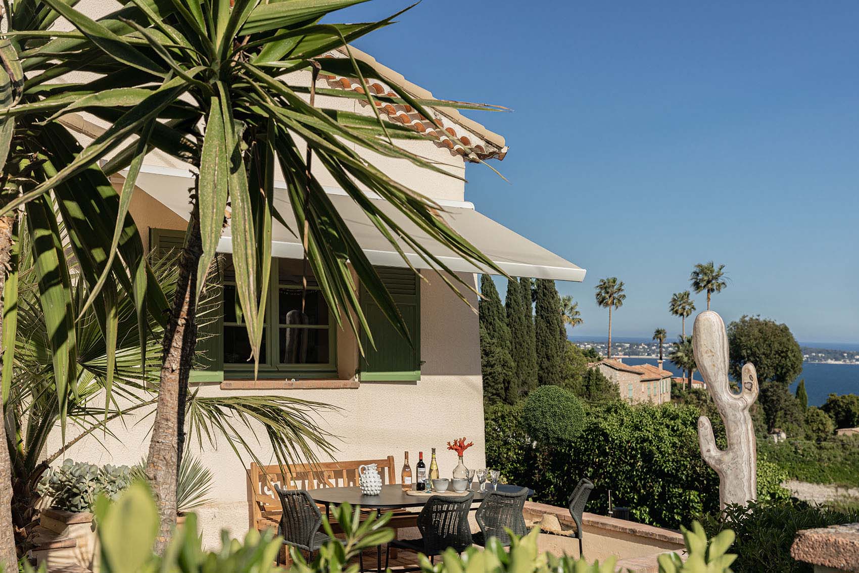 Provençal facade with green shutters, awning, palm tree, cactus sculpture and sea beyond